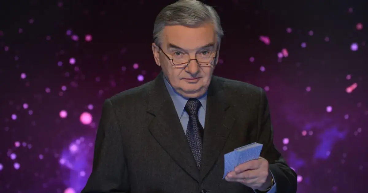 Tadeusz Sznuk w garniturze i okularach trzyma w ręku niebieskie karty na tle fioletowej, rozmytej scenografii.