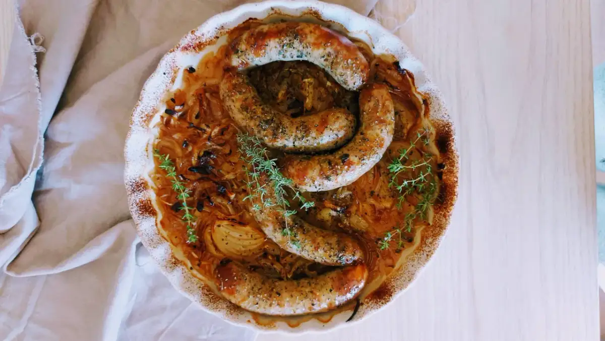 Złocista biała kiełbasa w piwie, zapiekana z cebulą i tymiankiem, prezentuje się apetycznie w ceramicznym naczyniu.