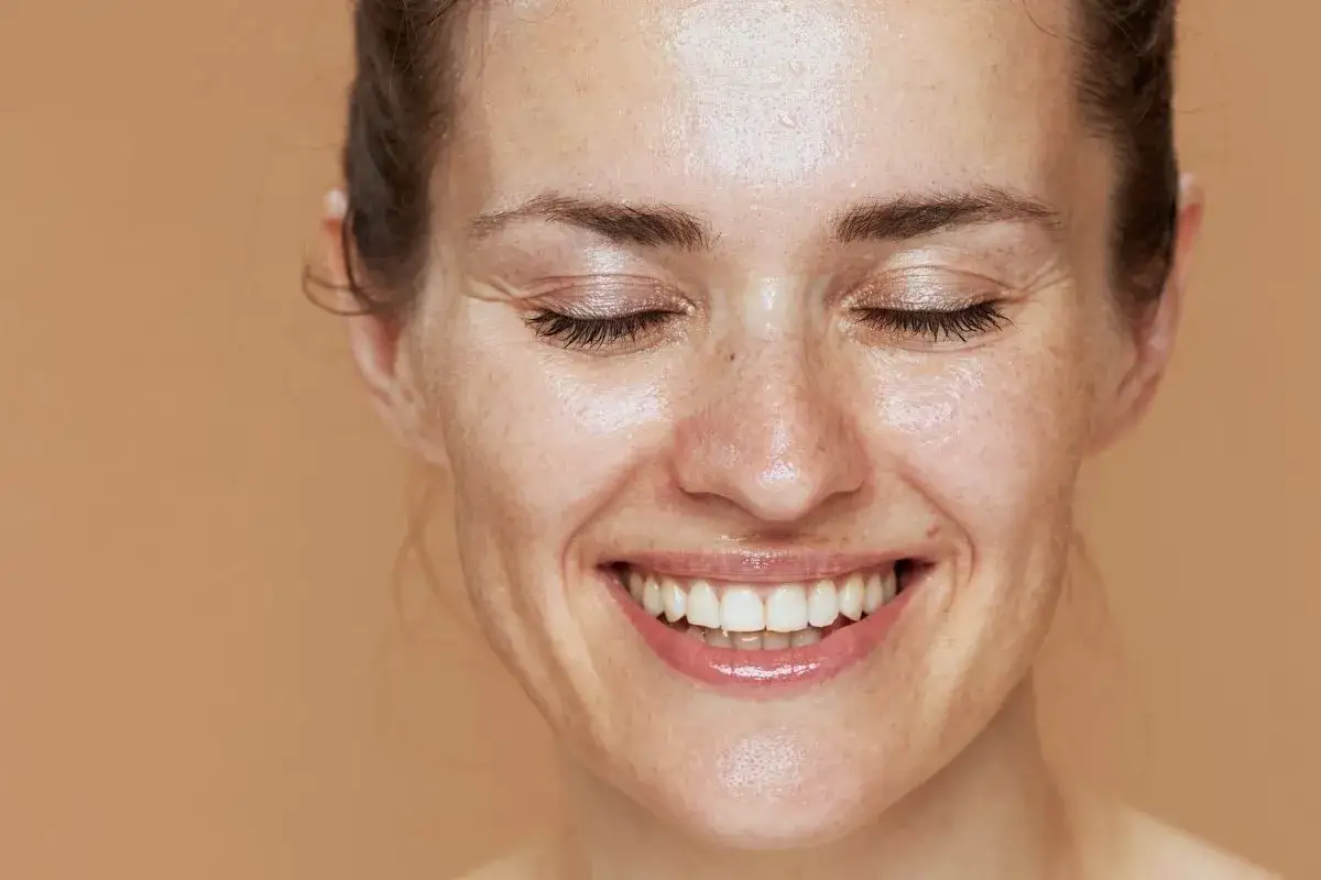 Uśmiechnięta kobieta z zamkniętymi oczami, jej skóra lśni po dwuetapowym oczyszczaniu twarzy.