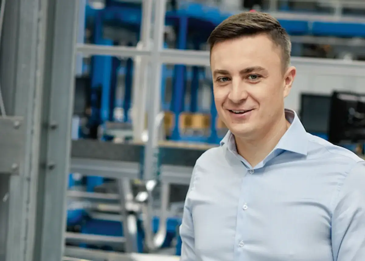 Uśmiechnięty mężczyzna w jasnoniebieskiej koszuli, na tle hali produkcyjnej. To pracownik firmy X KOM, która zajmuje się technologiami.