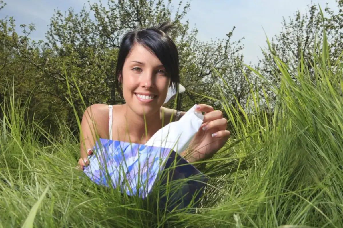 Uśmiechnięta kobieta w trawie, trzymająca pudełko chusteczek. Czas kiedy pyli lipa, a ona potrzebuje ulgi.