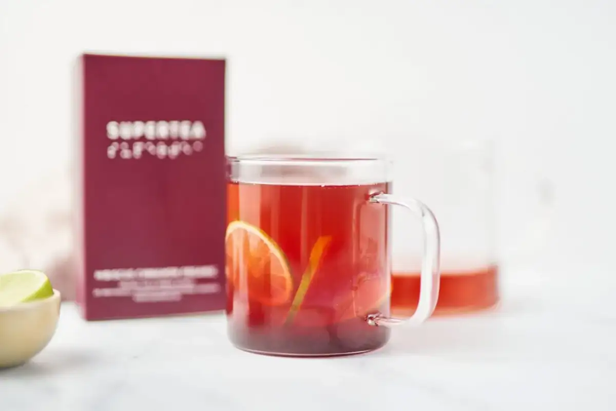 Szklanka z czerwonym napojem, plasterkami cytrusów i pudełkiem herbaty w tle.