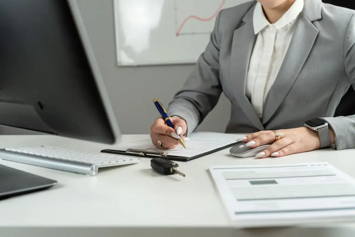 Kobieta w garniturze podpisuje dokumenty, obok kluczyki do auta i myszka. Zastanawia się, ile za km delegacji.