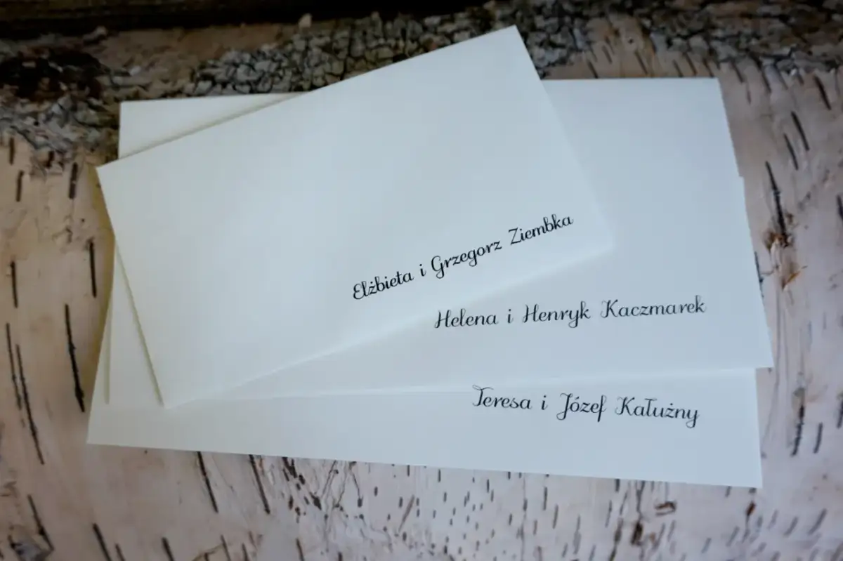 Białe koperty ślubne z elegancko wypisanymi nazwiskami gości na tle kory drzewa.