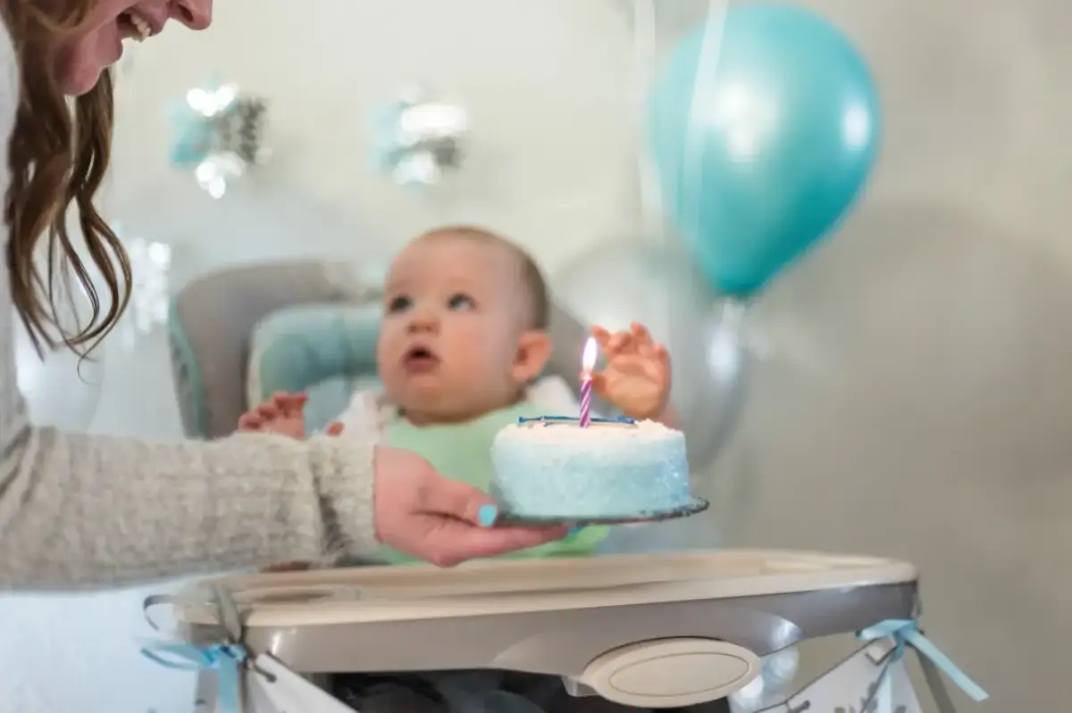 Mama podaje tort z jedną świeczką na roczek maluchowi. Balony i dekoracje tworzą radosną atmosferę.