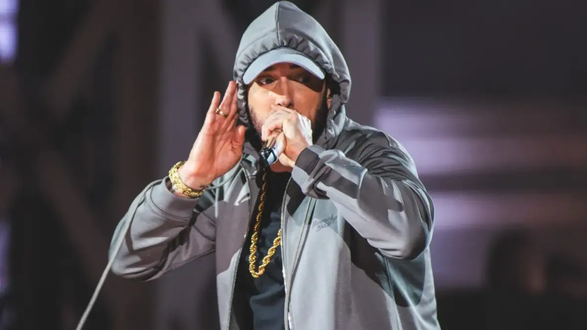 Eminem w szarej bluzie z kapturem i czapce, trzymający mikrofon i gestykulujący.