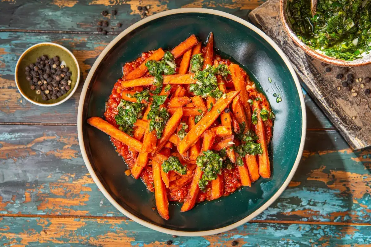 Pomarańczowe frytki z batatów podane z sosem pomidorowym i posypane zieloną, ziołową posypką. Obok miseczka z pieprzem.