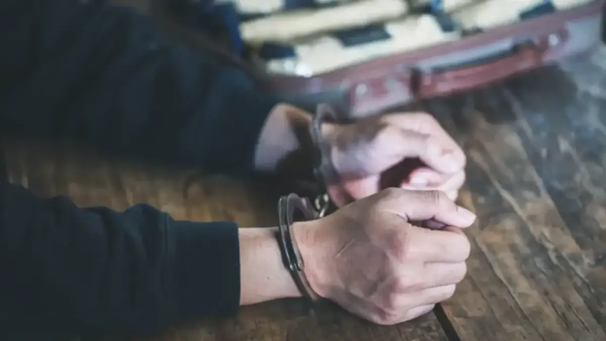 Ręce w kajdankach na drewnianym stole, w tle walizka z paczkami.