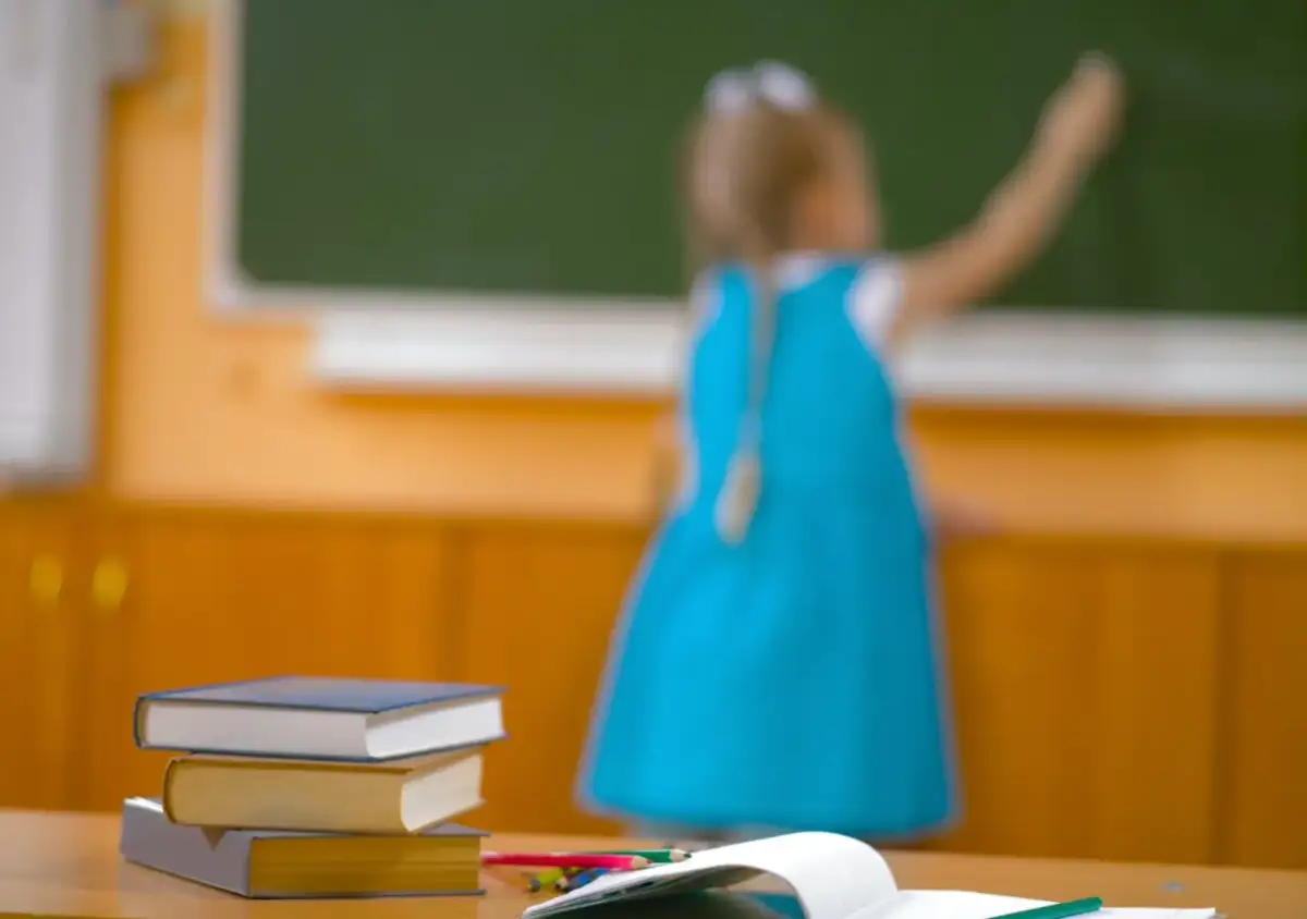 Dziewczynka w niebieskiej sukience pisze kredą po tablicy, a na pierwszym planie leżą książki. Jak to się ma do nauki zdalnej?