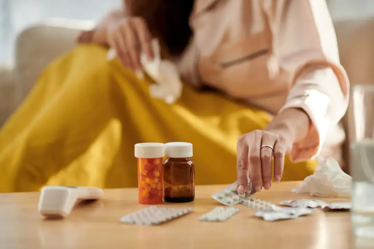 Chora osoba szuka leków na grypę przy nadciśnieniu. Na stole leżą termometr, butelki z lekami i blistry tabletek.