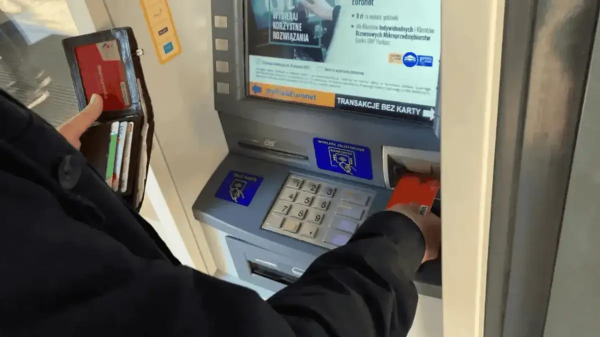 Osoba wkłada kartę do bankomatu, by wypłacić pieniądze. Widoczny portfel z kartami.