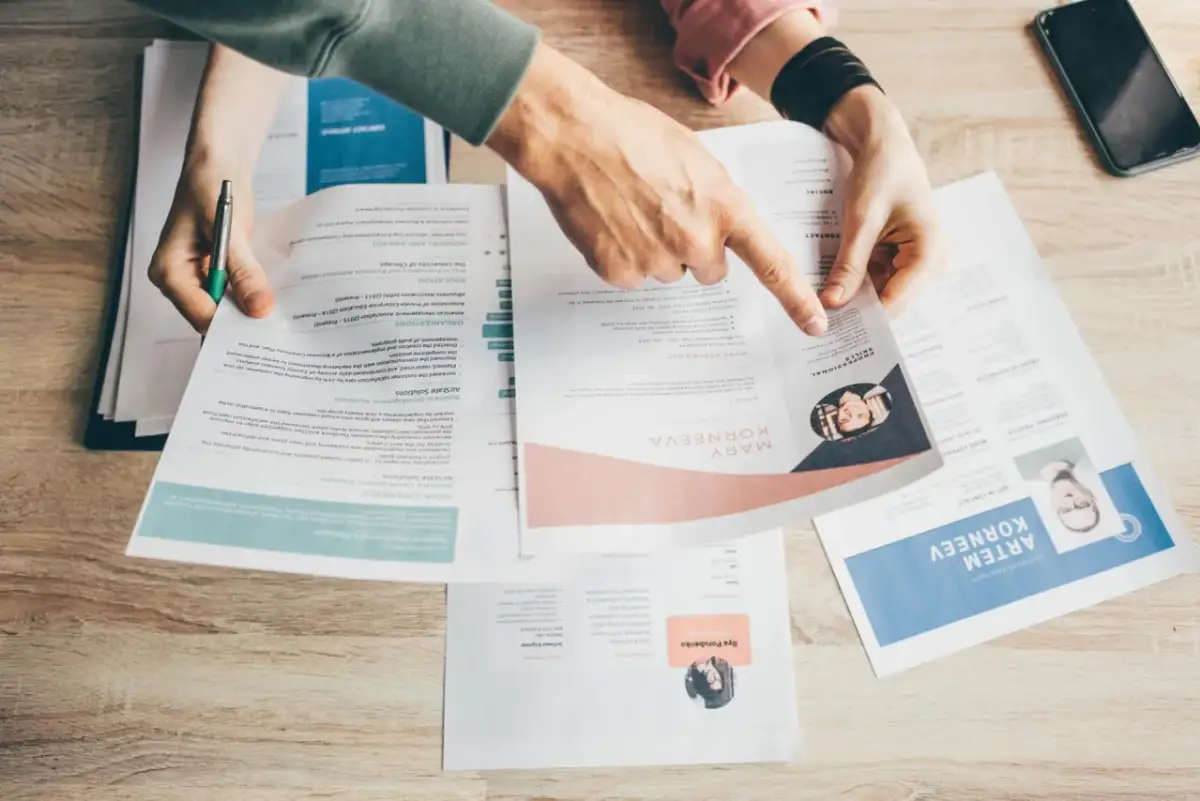 Dłonie przeglądające dokumenty z CV, na drewnianym tle.