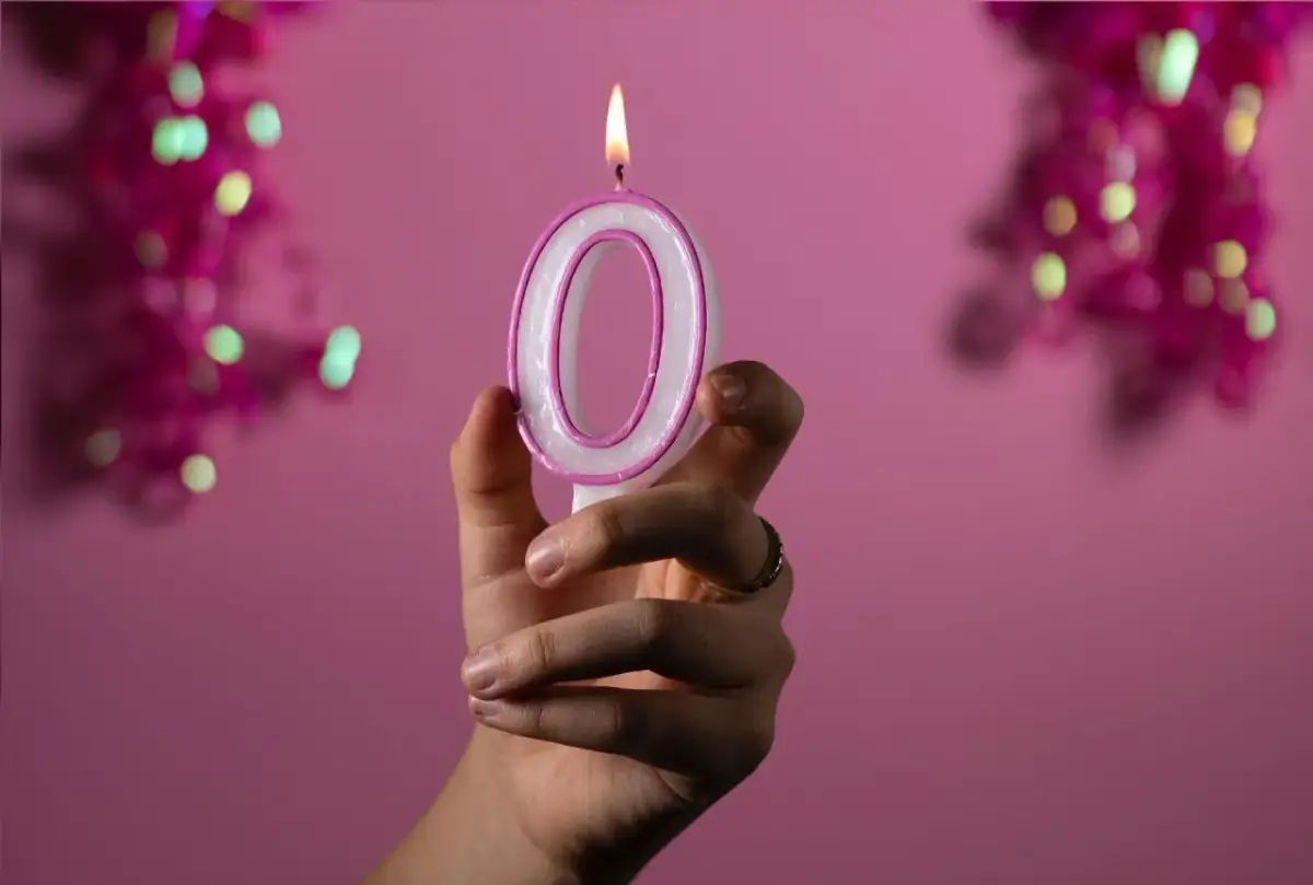 Dłoń trzyma zapaloną świeczkę w kształcie cyfry 0 na różowym tle.