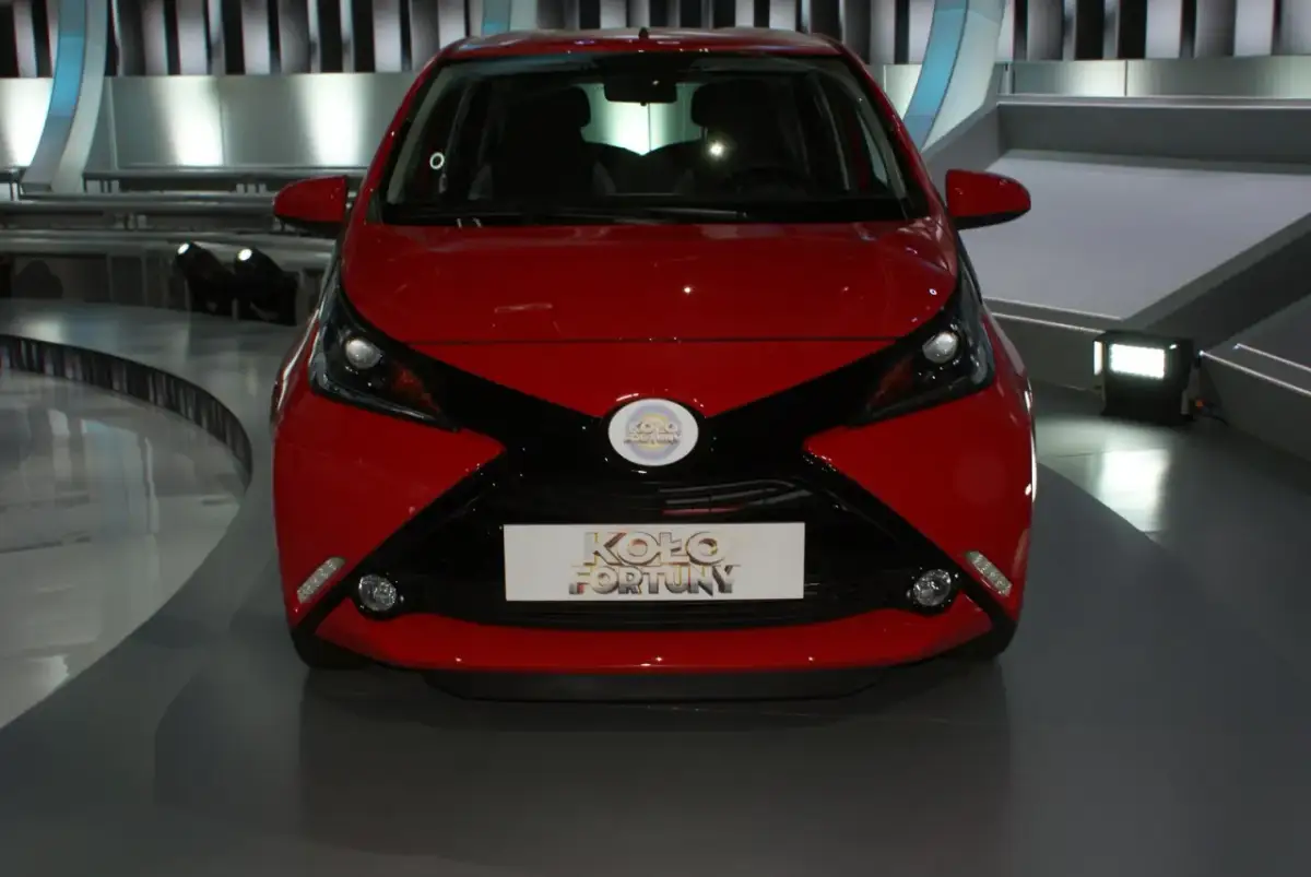 Czerwony samochód z czarnymi akcentami, prezentowany na scenie, z logo "Koło Fortuny" na masce.