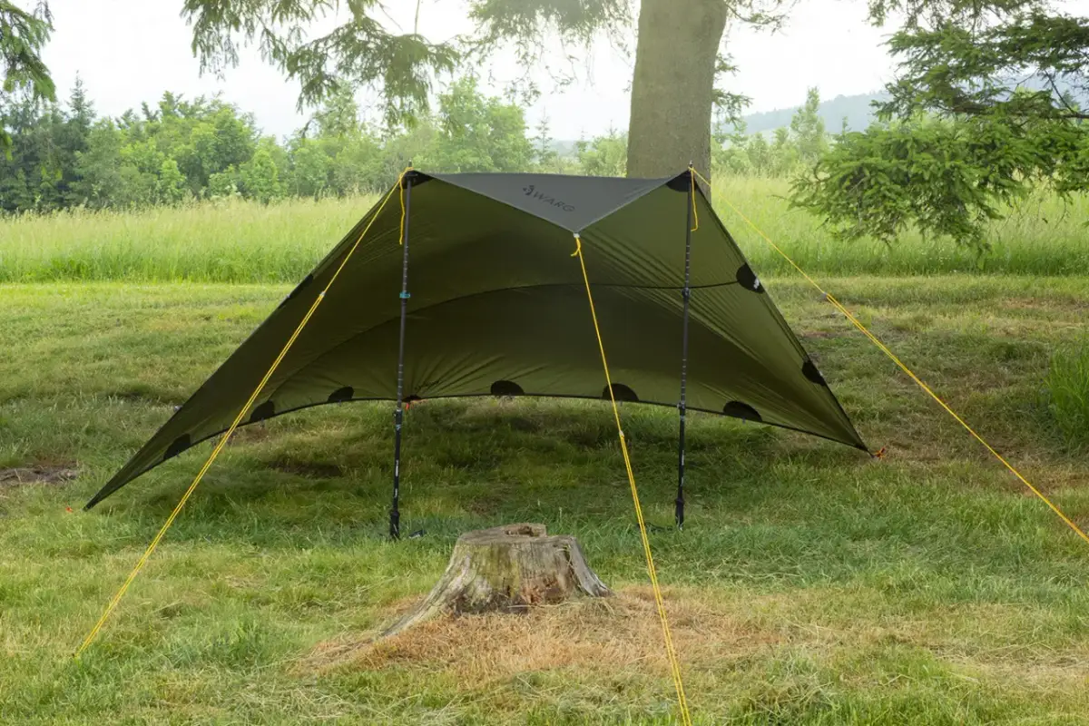 Zielony tarp rozstawiony na trawie, z żółtymi linkami. Pokazuje, jak zrobić namiot w prosty sposób, idealny na biwak.