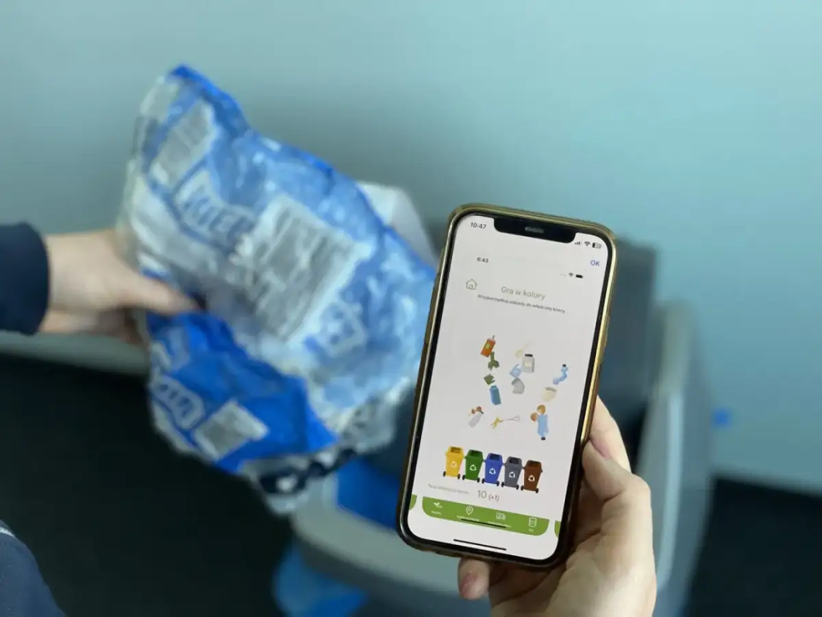 Aplikacja "jak segregować śmieci" pomaga w nauce sortowania odpadów. Na ekranie telefonu widać grę, gdzie trzeba dopasować przedmioty do kolorowych koszy.