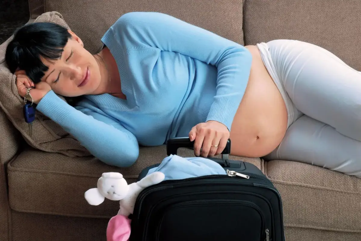 Zmęczona ciężarna kobieta śpi na kanapie, trzymając kluczyki do auta. Obok leży walizka z pluszową zabawką, gotowa na podróż, kiedy rodze.
