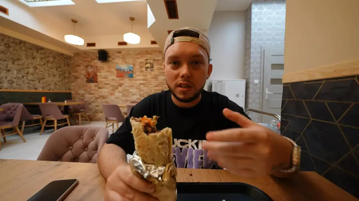 Mężczyzna w czapce z daszkiem trzyma w ręku dużego kebaba, zastanawiając się, gdzie jest najbliższy kebab.