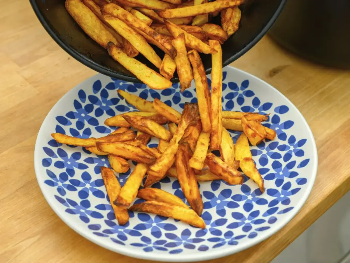 Chrupiące, złociste frytki wysypywane z czarnej patelni na biały talerz z niebieskimi kwiatami.