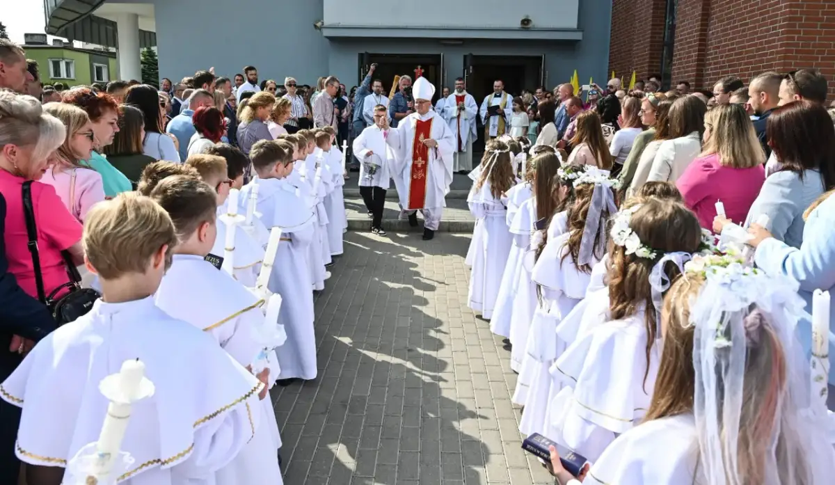 Dzieci w białych strojach komunijnych z zapalonymi świecami, biskup i księża przed kościołem.