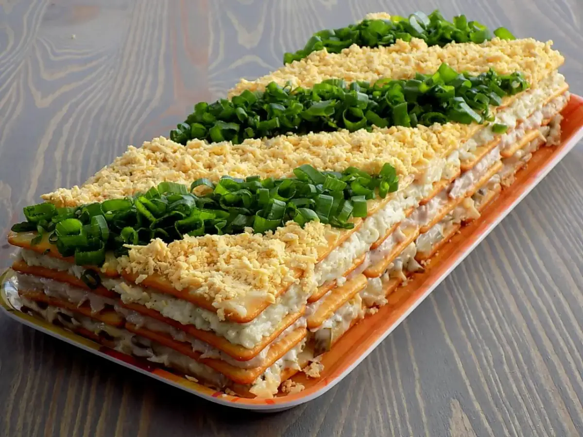 Warstwowa sałatka na krakersach, udekorowana startym jajkiem i szczypiorkiem, wygląda apetycznie.