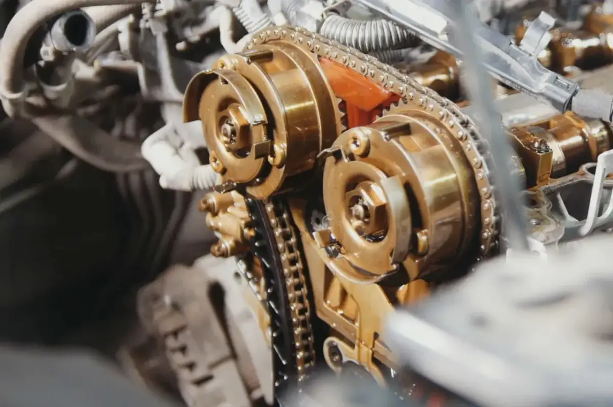 Zbliżenie na mechanizm silnika, gdzie trwa wymiana łańcucha rozrządu. Widoczne koła zębate i metalowy łańcuch pokryte olejem.