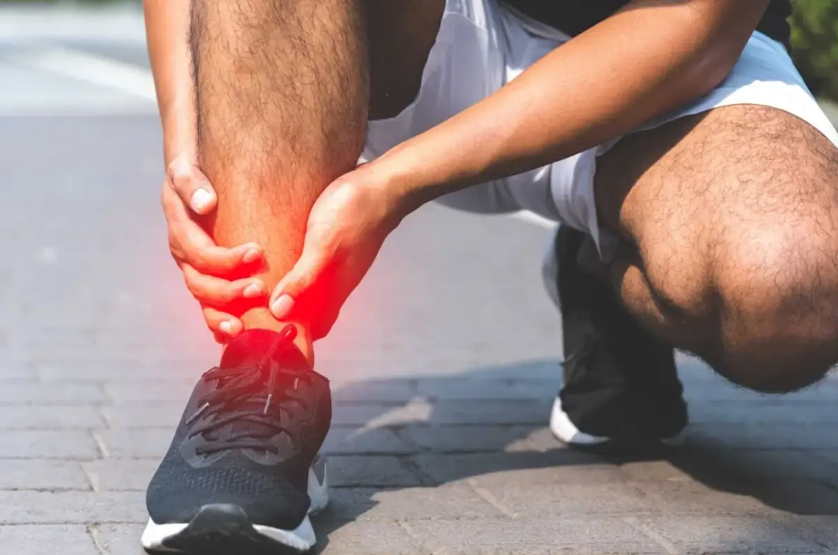 Osoba trzymająca się za bolesną, czerwoną kostkę w bucie sportowym.