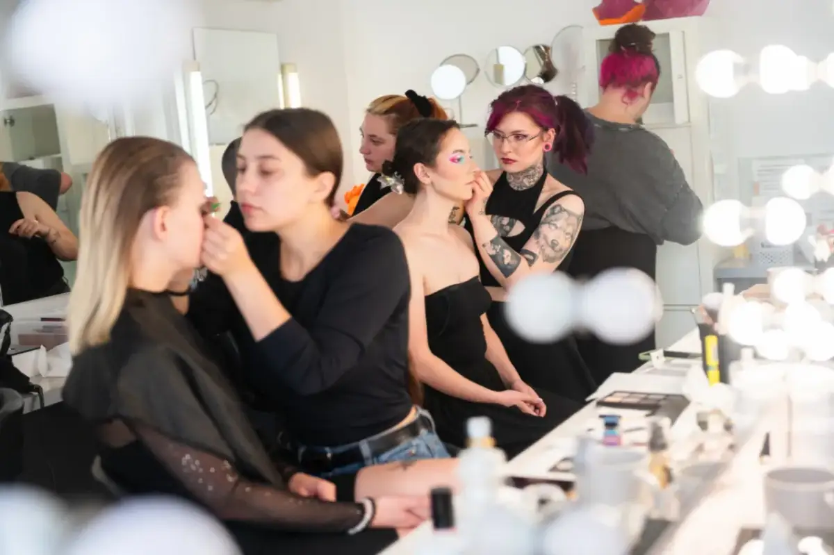 Charakteryzatorka filmowa pracuje nad makijażem modelki. W tle inne osoby przygotowują się do sesji.