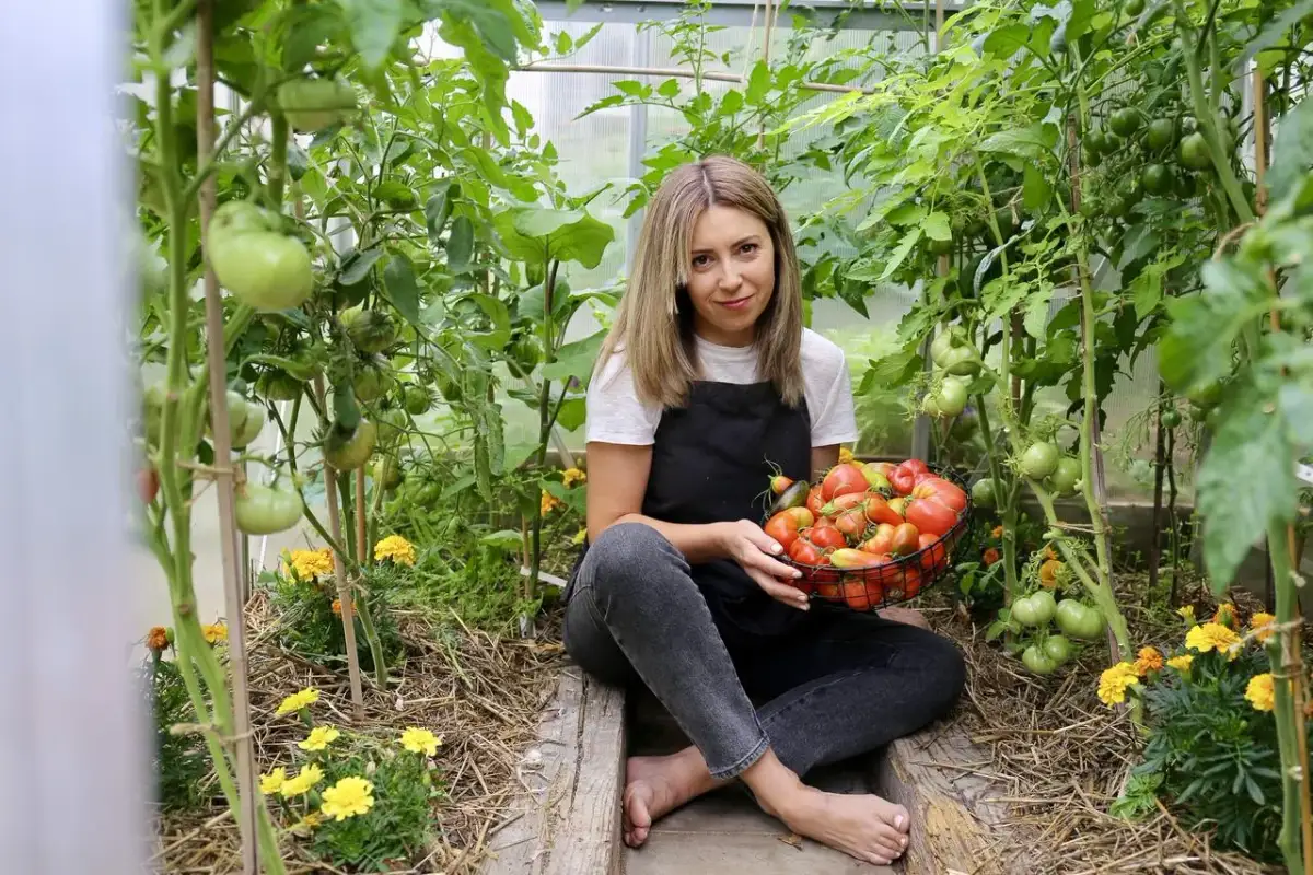 Kobieta w fartuchu siedzi w szklarni, trzymając koszyk pełen pomidorów. Wokół kwitną żółte kwiaty, a rośliny pomidorów pną się ku górze. To efekt udanego sadzenia warzyw.