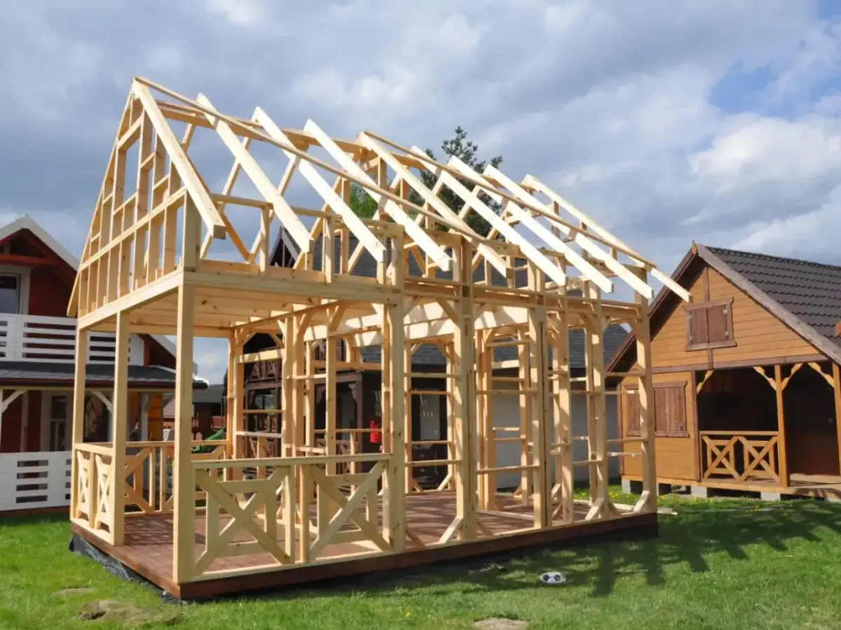 Drewniana konstrukcja domu szkieletowego w trakcie budowy na tle innych budynków.