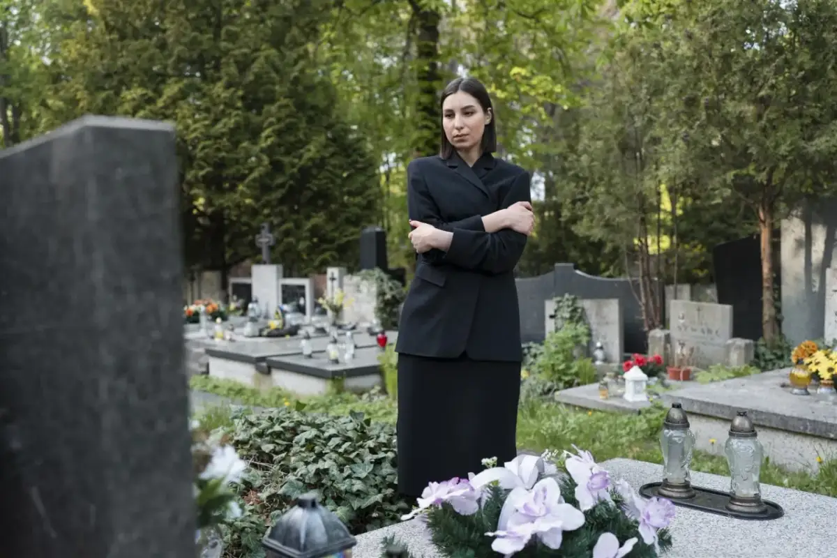 Smutna kobieta w czerni stoi na cmentarzu, otoczona nagrobkami. Jej postawa sugeruje częste chodzenie na cmentarz i głębokie przeżywanie żałoby.