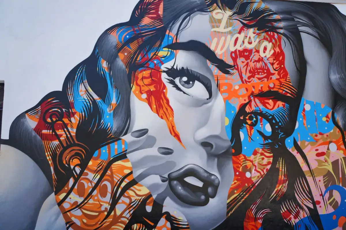 Mural z twarzą kobiety, pełen jaskrawych kolorów i abstrakcyjnych wzorów, oddaje dynamikę i wyrazistość cech pop artu.