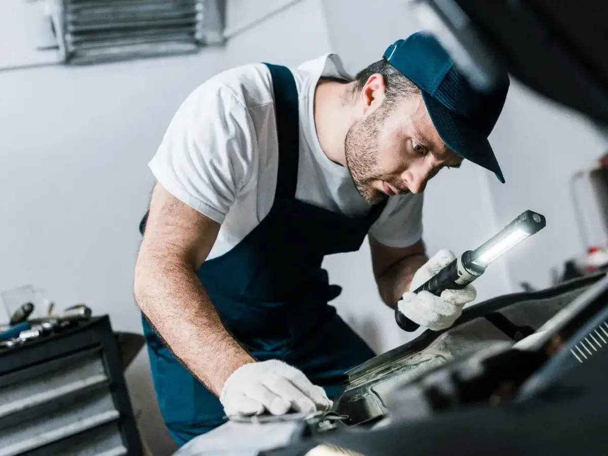 Mechanik w białej koszulce i granatowym kombinezonie, z latarką w ręku, sprawdza poziom płynu do sprzęgła w samochodzie.