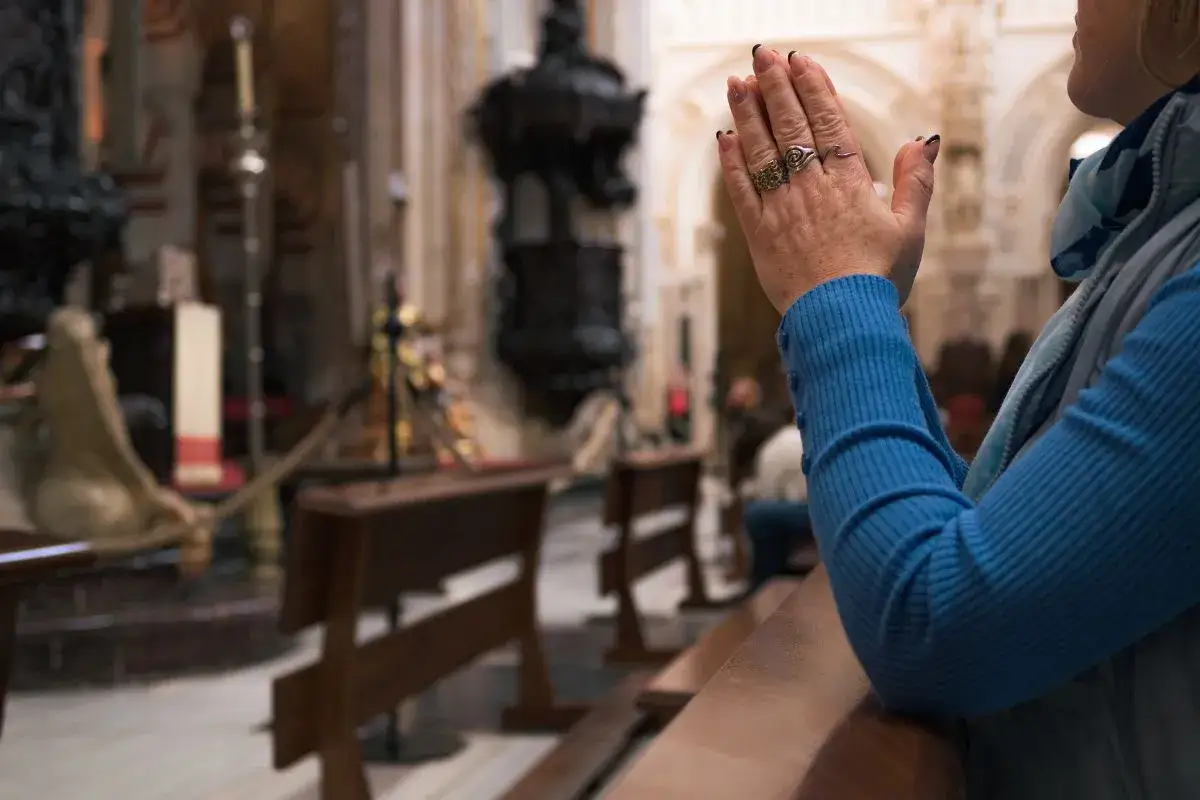 Dłoń kobiety w geście modlitwy w kościele, na tle ławek i ołtarza.