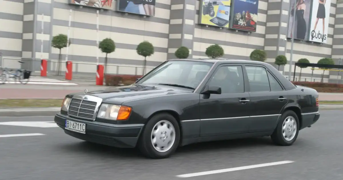 Ciemnoszary Mercedes W124 jedzie ulicą przed budynkiem z banerami.
