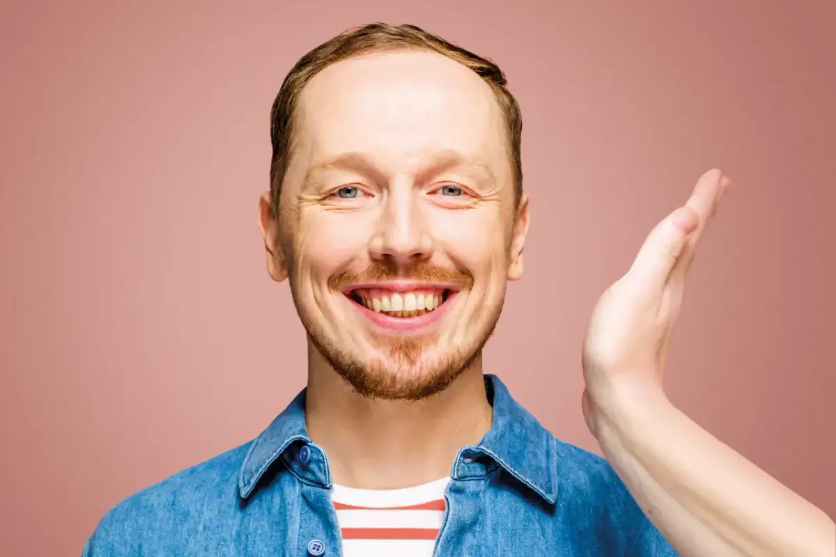 Uśmiechnięty mężczyzna w dżinsowej koszuli, jakby właśnie usłyszał świetny żart na występie syrek dąbrowski stand up.