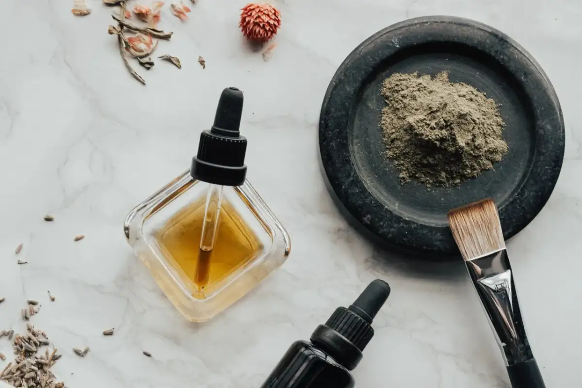 Składniki do domowego olejku eterycznego: suszone zioła, proszek w moździerzu, pędzelek i buteleczki z pipetą.