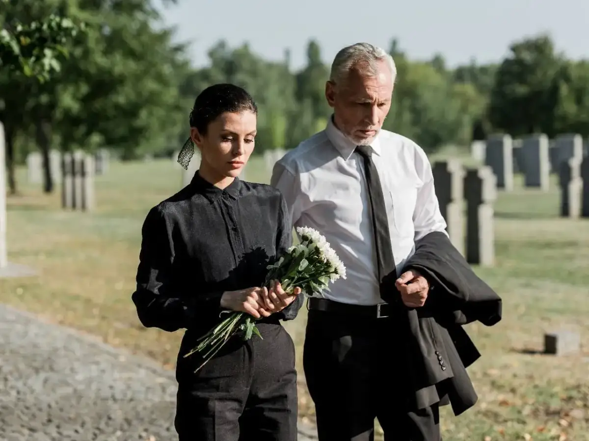 Para idąca przez cmentarz, kobieta w czarnej sukience trzyma bukiet białych kwiatów, mężczyzna w białej koszuli i czarnym krawacie niesie marynarkę.