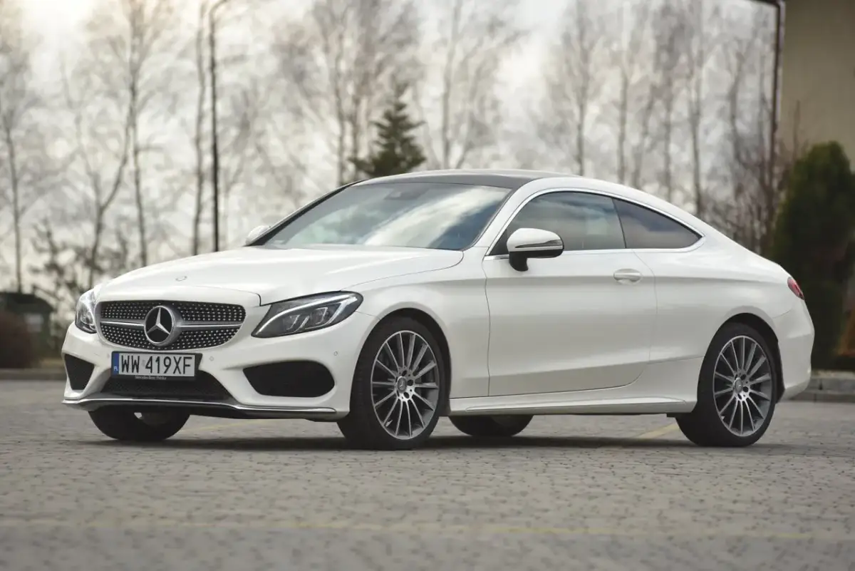 Biały Mercedes C klasa coupe z czarnym dachem, zaparkowany na brukowanej nawierzchni, z drzewami w tle.