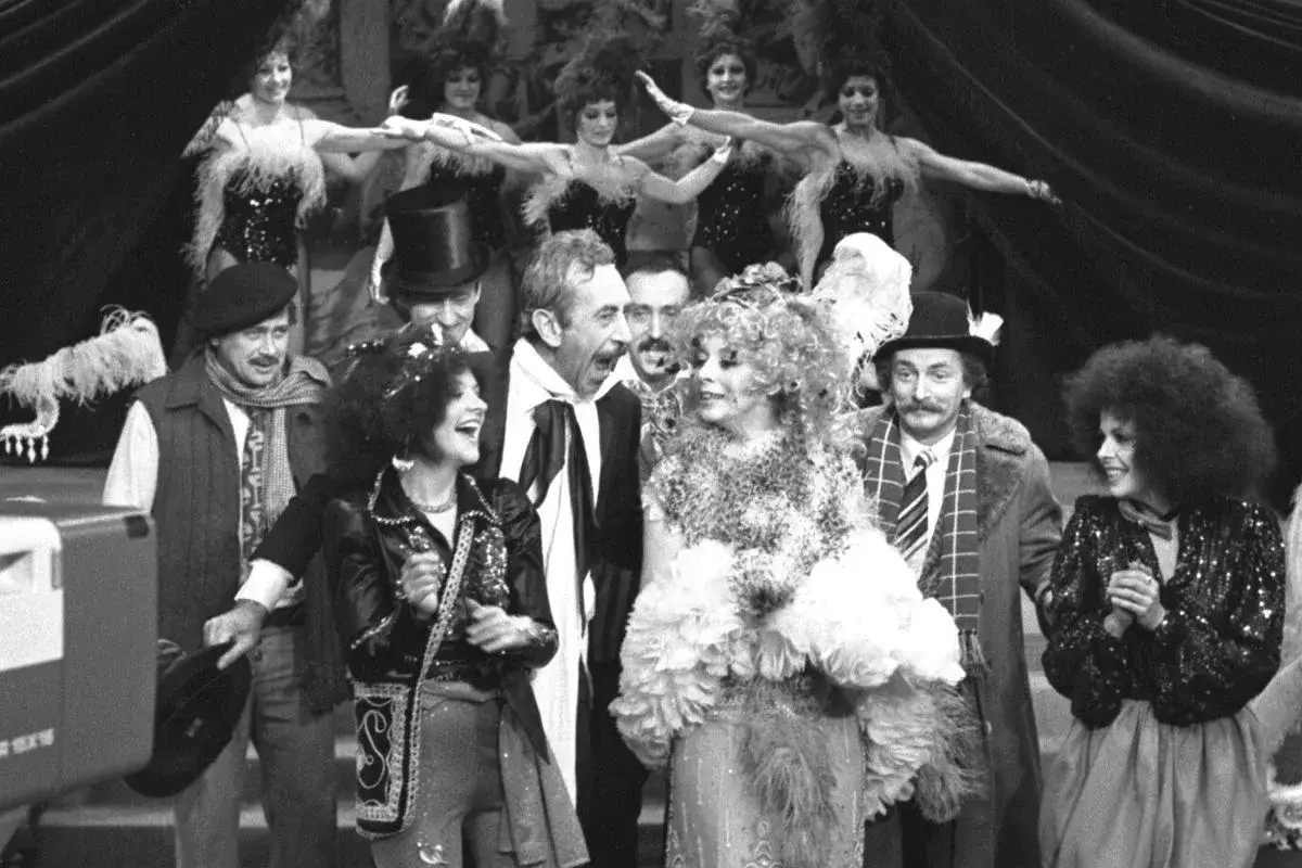 Artyści kabaretu Olgi Lipińskiej w barwnych strojach, wśród nich panie w piórach i panowie w kapeluszach, tworzą radosną scenę.