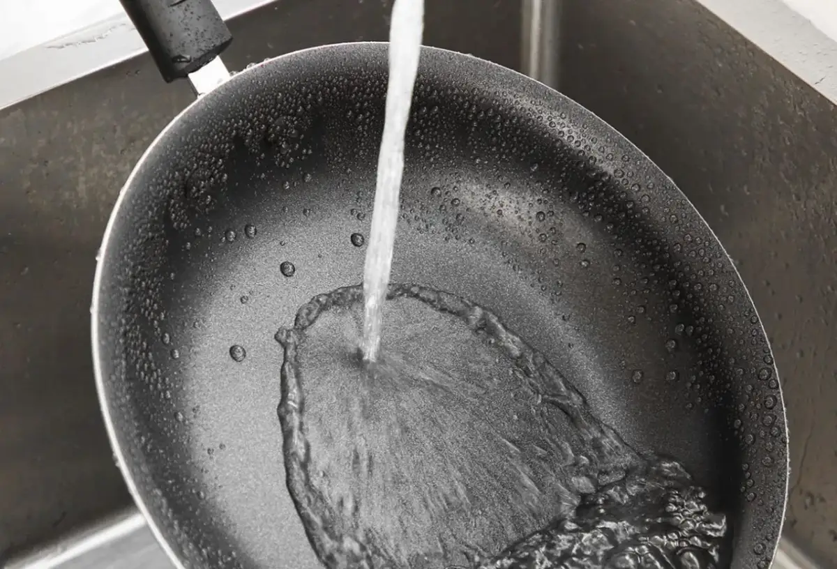 Czyszczenie patelni sodą i octem: strumień wody spływa po wewnętrznej stronie czarnej patelni w zlewie, tworząc pianę.