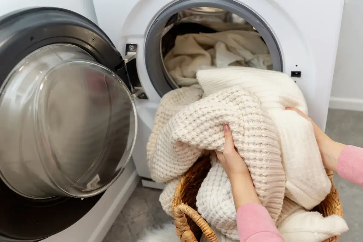 Ręce wkładają do pralki kosz z wełnianymi swetrami. Dowiedz się, jak prać wełnę w pralce, by zachować jej miękkość.