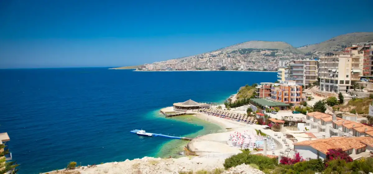 Widok na wybrzeże Sarandy, z turkusową wodą, plażą z parasolami i budynkami. W oddali widać panoramę miasta, przypominającą widok z Tirany.