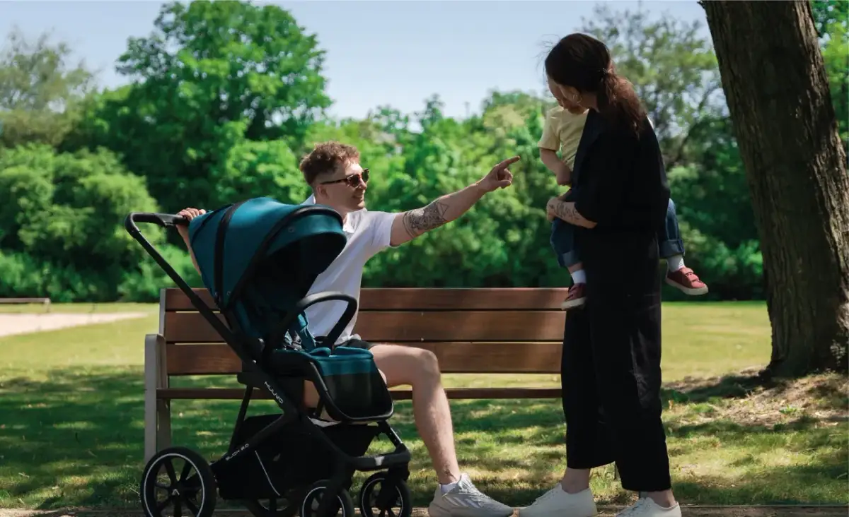 Rodzina na ławce w parku, ojciec wskazuje coś dziecku, zastanawiając się, kiedy zmienić gondolę na spacerówkę.
