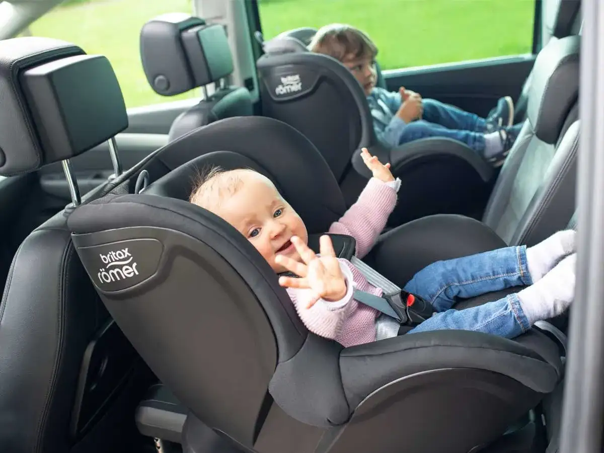 Uśmiechnięte niemowlę w foteliku samochodowym Britax Römer. Obok starsze dziecko. Bezpieczna podróż do jakiego wieku dziecko w foteliku jest kluczowa.