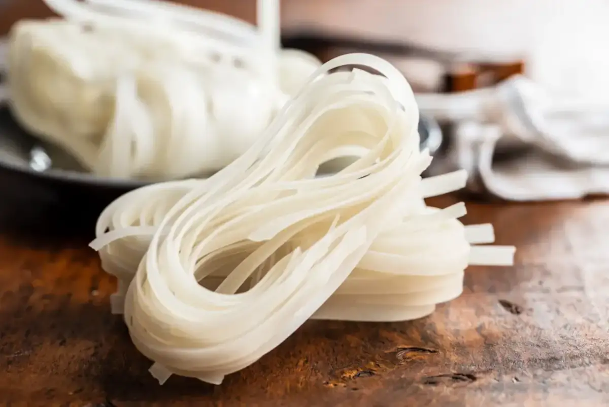 Stos suchych, białych wstążek makaronu ryżowego na drewnianym blacie.