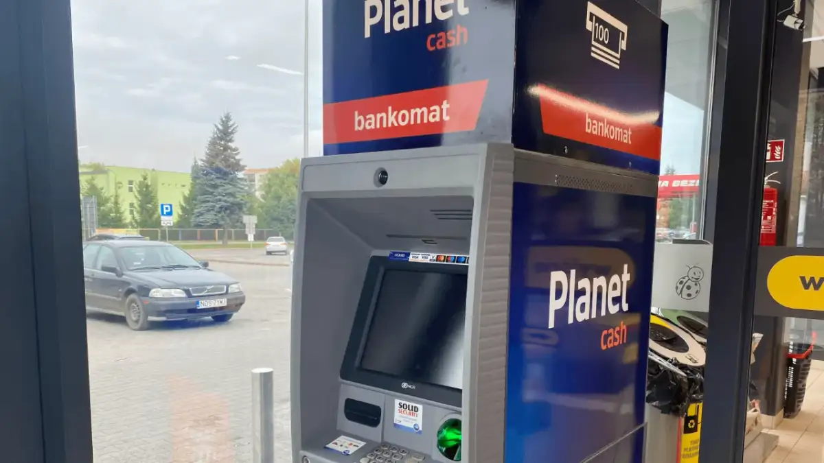 Bankomat Santander Planet Cash przy parkingu. Obok stoi czarny samochód.