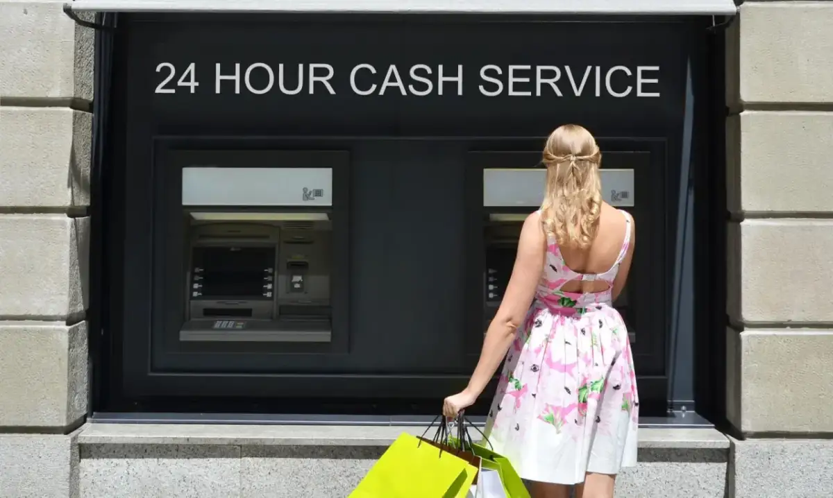 Kobieta w letniej sukience z zakupami przed bankomatem. Czy można wypłacić 20 zł z bankomatu PKO?