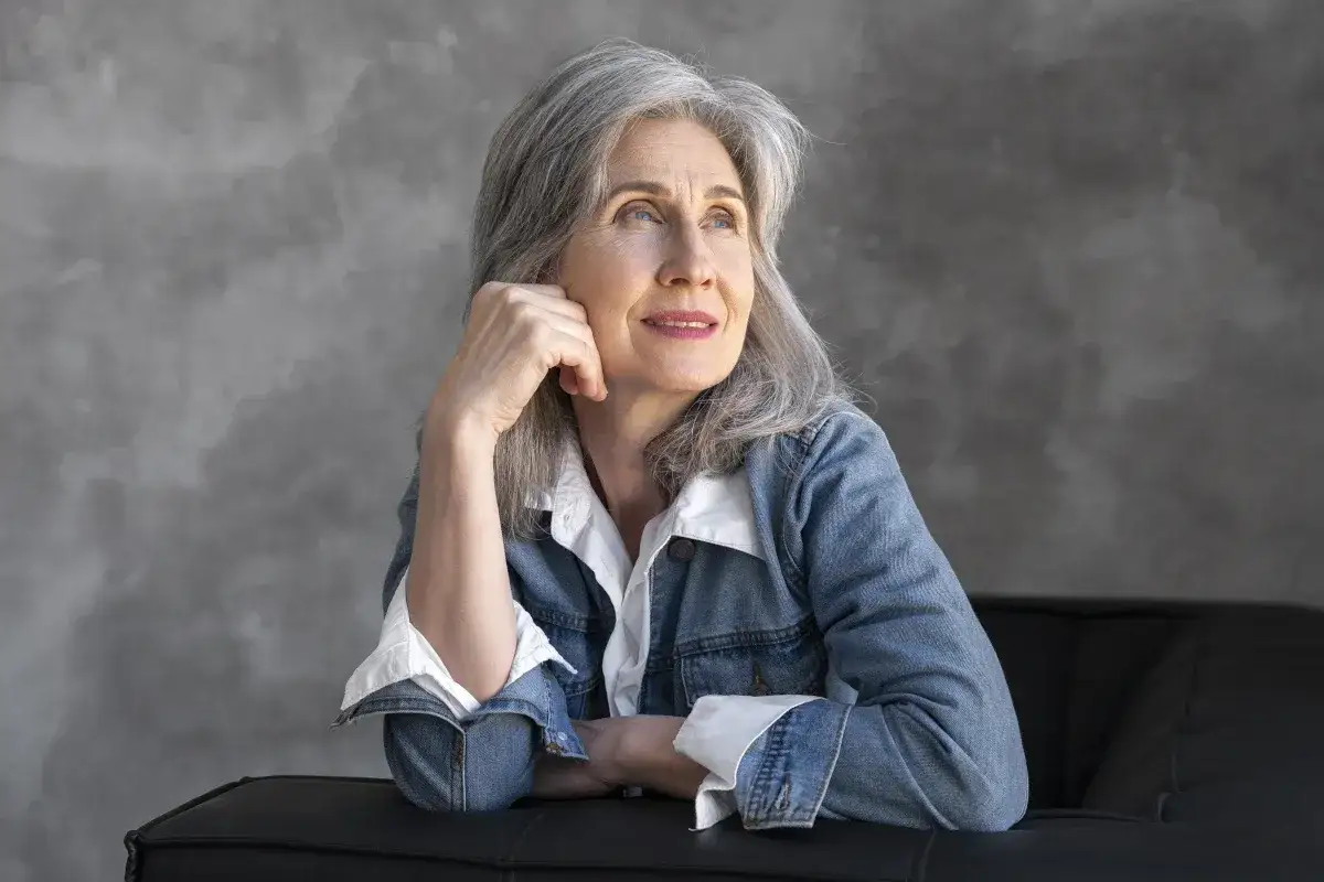 Kobieta z siwymi włosami, oparta o czarną kanapę, zamyślona. Może myśli o ziołach na pamięć, które pomagają jej zachować jasność umysłu.