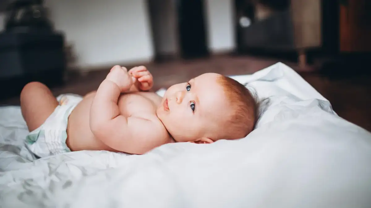 Niemowlę w pieluszce leży na białym prześcieradle, patrząc w bok.